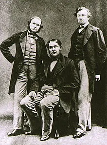 Three middle-aged men, with the one in the middle sitting down. All wear long jackets, and the shorter man on the left has a beard.